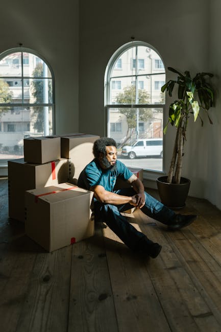 A man with curly hair and a beard, dressed in a teal shirt and dark jeans, is sitting on a wooden floor inside a room with large, arched windows. He is surrounded by several medium-sized cardboard boxes, some sealed with red tape, which are stacked around him and positioned on the floor. Natural light illuminates the space through the windows, revealing a view of a street with parked cars and nearby buildings. A tall potted plant with broad green leaves is situated near the window on the right side, adding greenery to the interior. The scene depicts a moment during a home relocation process, where the man appears to be taking a break amidst packing and moving activities. This image is related to house removals and furniture transport services, such as those provided by Man and Van Crayford, supporting packing, loading, and transportation tasks involved in a house move.