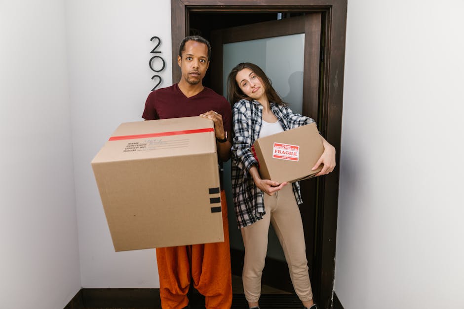A man and a woman stand inside a doorway of a residential property, each holding a cardboard moving box. The man, dressed in a maroon shirt and orange trousers, has a serious expression and is holding a large box with a red and white label, while the woman, wearing a checkered shirt and beige trousers, leans slightly to the side with a slight smile and holds a smaller box marked with a red label reading 'FRAGILE.' The background shows a dark wooden doorframe and white walls, indicating an interior space during a home relocation process. The scene suggests the packing and loading phase of furniture transport and household items, with visible cardboard boxes and minimal packaging materials balancing a professional yet casual environment. The overall setting reflects moving logistics managed by a removal company, with the process of loading household items for transport in progress, consistent with services offered by Man and Van Crayford at [PAGE_TITLE].