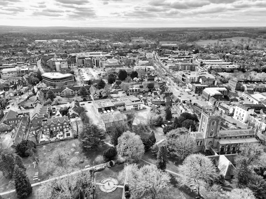 An aerial black-and-white photograph captures a small town with a mix of residential, commercial, and institutional buildings, including a prominent church with a tall steeple situated amidst a canopy of trees. The urban layout features streets and roads radiating outward, with vehicles visible parked along the curbs and moving through intersections. In the foreground, a park area is visible with pathways, benches, and numerous deciduous and evergreen trees, some with bare branches indicating a season of late autumn or winter. Adjacent to the park, there are houses and small apartment buildings, some with pitched roofs and gardens. The background reveals a more densely built central business district with modern and traditional structures, leading into open countryside with fields and woodlands on the horizon. The scene illustrates the complex environment involved in house removals and home relocation logistics, with Man and Van Crayford positioned as a professional provider of removals services that facilitate efficient furniture transport and packing processes.