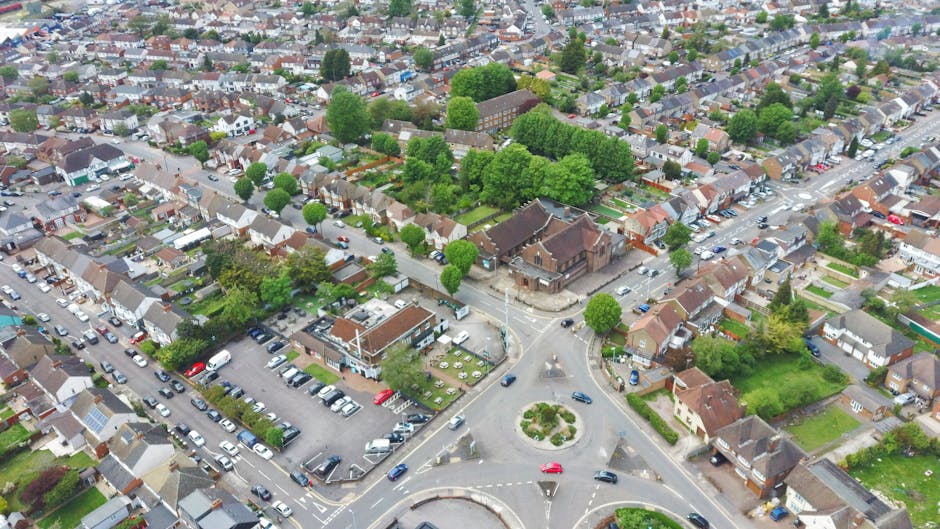 An aerial view of a residential neighbourhood in Crayford showing a roundabout at the intersection of multiple small streets lined with detached and terraced houses, many with front gardens and trees. In the foreground, a parking lot filled with cars is visible, adjacent to a brick building, possibly a community centre or local facility. The area is surrounded by greenery, including mature trees and patches of grass, indicating a well-established suburban community. The scene is captured during daylight, with clear weather and natural light illuminating the scene. The image reflects the typical urban environment where home removals and furniture transport by Man and Van Crayford could take place, especially near busy roads and parking areas suitable for loading and unloading household goods during the packing and moving process.