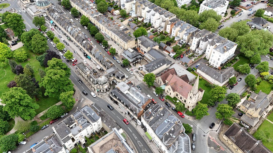 An aerial black-and-white photograph captures a small town with a mix of residential, commercial, and institutional buildings, including a prominent church with a tall steeple situated amidst a canopy of trees. The urban layout features streets and roads radiating outward, with vehicles visible parked along the curbs and moving through intersections. In the foreground, a park area is visible with pathways, benches, and numerous deciduous and evergreen trees, some with bare branches indicating a season of late autumn or winter. Adjacent to the park, there are houses and small apartment buildings, some with pitched roofs and gardens. The background reveals a more densely built central business district with modern and traditional structures, leading into open countryside with fields and woodlands on the horizon. The scene illustrates the complex environment involved in house removals and home relocation logistics, with Man and Van Crayford positioned as a professional provider of removals services that facilitate efficient furniture transport and packing processes.