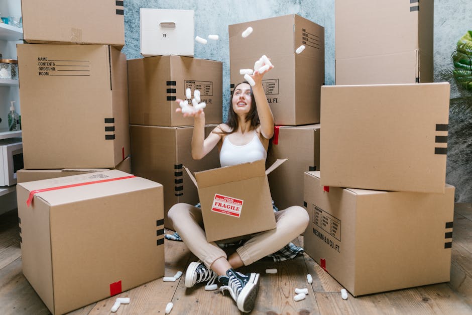 A young woman with long dark hair, wearing a white sleeveless top and beige pants, is sitting cross-legged on a light wooden floor surrounded by multiple large cardboard moving boxes, some with red and black tape and labels. She is smiling and looking up as she throws pieces of white packing foam into the air, which are scattered around her and some are falling from her hands. The boxes are stacked and arranged in the background inside a room with light blue textured walls, a small white side table holding a glass bottle, and a green leafy plant in the corner. The scene captures a playful moment during home relocation, illustrating the packing process involved before house removals, with the presence of moving equipment such as the foam packing material supporting the logistics of furniture transport and packing and moving services provided by Man and Van Crayford.