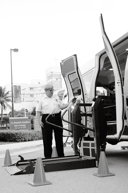 A man wearing glasses and a white shirt stands next to a white van with open side and rear doors, involved in a home relocation process. The van is parked on a paved street near residential buildings, with a small set of foldable ramps and orange traffic cones positioned at the vehicle's entry point to facilitate wheelchair or trolley access. Inside the van, visible black storage compartments and a wheelchair lift system are being used to load or unload furniture or boxes. The surrounding environment includes street lamps, a palm tree, and signage indicating nearby parking facilities. The image captures the loading process during a house removal, with the man preparing equipment or overseeing the packing and transport of household belongings, reflecting professional furniture transport and packing and moving activities often managed by services like Man and Van Crayford.