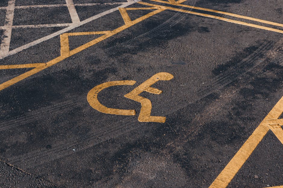 A close-up view of a designated accessible parking space on an asphalt surface, featuring a yellow painted wheelchair symbol in the center, surrounded by yellow diagonal lines indicating a no-parking zone. The surface shows slight wear and tire marks, with the parking area situated near a building entrance, which is partially visible in the background. The space is empty, ready for vehicle parking associated with house removals or relocation services. This parking area is part of a commercial or residential location, with the space allocated for ease of access during furniture transport, packing, and moving activities handled by Man and Van Crayford, a company specialising in removals and home relocation services.