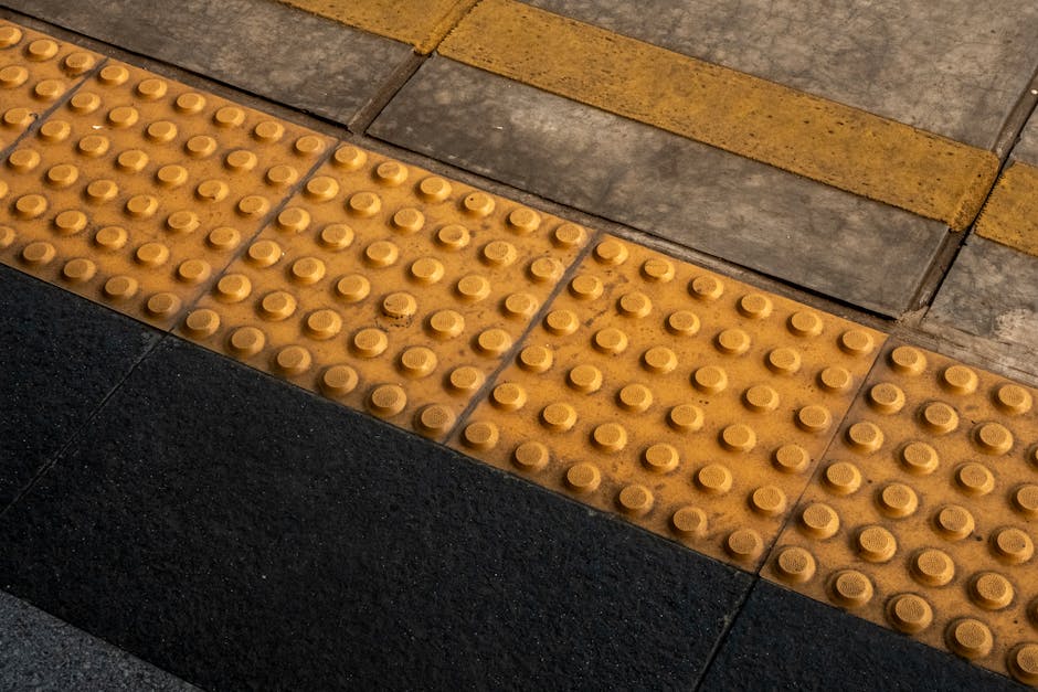 Close-up view of a tactile paving surface on a pavement, featuring yellow raised domes arranged in a grid pattern to assist visually impaired pedestrians. The paving overlaps a section of dark grey concrete, with a yellow painted line running parallel along the top edge of the image, indicating a boundary or guide for safe crossing areas. The tactile tiles are adjacent to standard concrete slabs with metal expansion joints visible between the sections. The area appears to be part of an urban street scene, possibly near a building entrance or a designated walkway, with natural lighting highlighting the textures and materials. This type of paving is often used during home relocation or furniture transport to facilitate safe movement for pedestrians. The image may be linked with house removals or moving services, as provided by companies such as Man and Van Crayford, who assist with packing, loading, and transporting items through such accessible pathways.