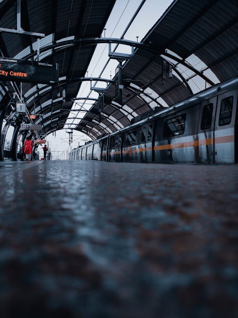 Interior view of Crayford railway station platform during daytime, showing a train with a silver exterior and orange stripe parked alongside the platform. The station features a curved, arched roof made of metal and glass panels allowing natural light to illuminate the area. The platform surface is textured, with a low angle perspective highlighting the details of the textured flooring. A few passengers, some with luggage and backpacks, are visible near the platform edge, waiting or preparing to board the train. Overhead signs and digital displays are mounted under the roof, providing travel information. The scene captures the typical environment for home relocation or furniture transport operations involving station-based logistics, as managed by Man and Van Crayford, a professional removals service specializing in local moving solutions.
