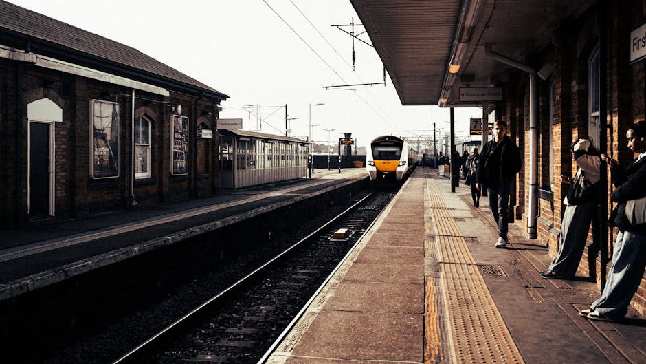 A view of Crayford railway station platform taken during daylight hours, showing a yellow and white train arriving or departing in the background. The platform is lined with tactile paving strips near the edge for safety, and has a brick wall on one side with some windows and posters. Several passengers are waiting on the platform, standing or leaning against the wall, dressed in casual clothing. The station has an overhanging roof supported by metal poles, providing shelter to waiting passengers. The scene captures the typical environment of a busy local station, with ambient lighting highlighting the details of the platform, train, and waiting people, reflecting aspects of home relocation or travel logistics associated with moving services from Man and Van Crayford.