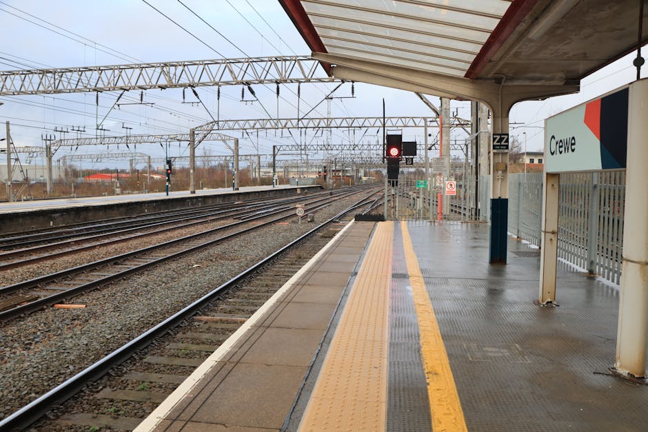 Interior view of Crayford railway station platform during daytime, showing a train with a silver exterior and orange stripe parked alongside the platform. The station features a curved, arched roof made of metal and glass panels allowing natural light to illuminate the area. The platform surface is textured, with a low angle perspective highlighting the details of the textured flooring. A few passengers, some with luggage and backpacks, are visible near the platform edge, waiting or preparing to board the train. Overhead signs and digital displays are mounted under the roof, providing travel information. The scene captures the typical environment for home relocation or furniture transport operations involving station-based logistics, as managed by Man and Van Crayford, a professional removals service specializing in local moving solutions.
