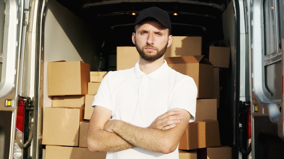 A man wearing a black cap and white polo shirt is standing with his arms crossed inside a van during the loading process for a house removal service. The van's interior is filled with stacked cardboar