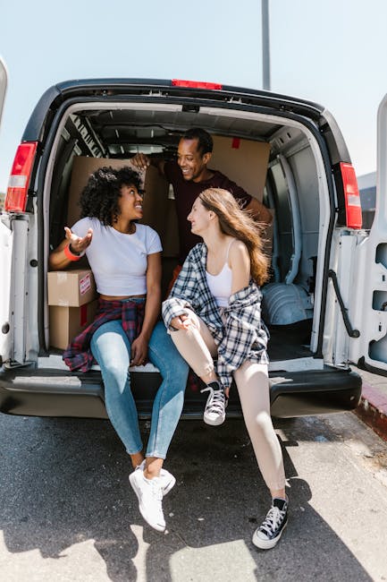 A man wearing a black cap and white polo shirt is standing with his arms crossed inside a van during the loading process for a house removal service. The van's interior is filled with stacked cardboar