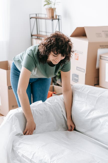 A woman with curly brown hair, dressed in a green T-shirt and blue jeans, is carefully wrapping or unwrapping furniture with protective white plastic or fabric, indicating a home relocation process. S