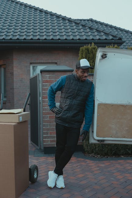 A man wearing casual clothing, including a white t-shirt, jeans, and a black cap, is carrying three medium-sized cardboard boxes toward a white panel van parked on a suburban street. The van’s rear 