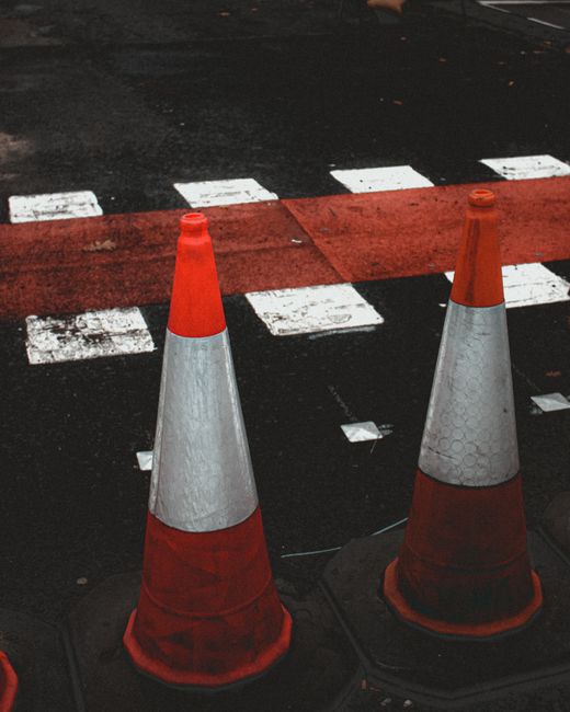 Two orange and white traffic cones placed on a paved road next to a red and white striped pedestrian crossing, with some worn white markings visible on the dark asphalt. The cones are positioned to restrict access or indicate a temporary closure, likely related to home relocation or moving vehicle parking controls in Crayford. In the background, a red painted area on the road and partial white lines are visible, suggesting a designated loading zone or restricted parking area, relevant to house removals. This scene illustrates the necessary traffic management measures, such as permits and road closure rules, for professional furniture transport and packing and moving services provided by Man and Van Crayford, ensuring compliance during the logistics process.