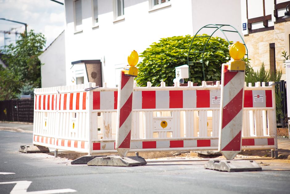 A road closure barrier positioned on a residential street, consisting of a white plastic fence with red reflective stripes, supported by black rubber bases for stability. The barrier features two vertical red and white striped poles with yellow warning lights on top, connected by a curved metal arch. A white informational sign is attached to the fence. Behind the barrier, there is a house with white exterior walls, multiple windows, and a stone-clad section near the entrance. A large green bush and some smaller plants are visible in front of the house. The pavement in front of the barrier appears to be under partial maintenance or construction, with a section of the road closed off for safety, reflecting local road closure rules affecting house removals and moving trucks. This setup is associated with move logistics and transport restrictions in Crayford, as managed by [COMPANY_NAME].