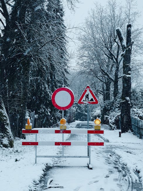Two orange and white traffic cones placed on a paved road next to a red and white striped pedestrian crossing, with some worn white markings visible on the dark asphalt. The cones are positioned to restrict access or indicate a temporary closure, likely related to home relocation or moving vehicle parking controls in Crayford. In the background, a red painted area on the road and partial white lines are visible, suggesting a designated loading zone or restricted parking area, relevant to house removals. This scene illustrates the necessary traffic management measures, such as permits and road closure rules, for professional furniture transport and packing and moving services provided by Man and Van Crayford, ensuring compliance during the logistics process.