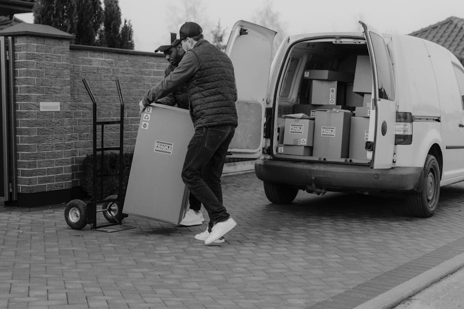 Two men are engaged in a home relocation process on a paved driveway, unloading a cardboard box with a 'Fragile' warning label from the back of a white delivery van. One man, dressed in a cap and dark jacket, is lifting the box onto a small, hand-pushed trolley, while the other, wearing glasses and a puffer jacket, assists by steadying the box. Inside the open van, there are several other cardboard boxes of various sizes, some sealed with packing tape, stacked on the vehicle's floor, indicating organised packing and furniture transport for a house move. The scene takes place outdoors near a low brick wall and a gated entrance, with natural light providing clear visibility, reflecting typical conditions during a same-day removal service. This image illustrates the logistical process of loading household items, involving careful packing materials and equipment like the trolley, as part of professional removals offered by Man and Van Crayford, focusing on efficient and secure moving and packing for house relocations.