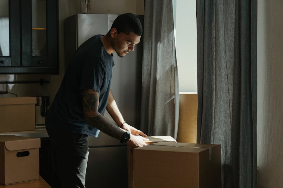 A man with dark skin and short black hair, wearing a dark T-shirt and jeans, is inside a residential kitchen preparing for a house move. He is leaning over a cardboard box, which is placed on a wooden surface, possibly packing or preparing items for transportation. The kitchen features dark cabinets and a stainless steel refrigerator visible in the background. Natural light is streaming through a window beside him, partially covered by grey curtains, illuminating the scene. Several other cardboard boxes are arranged nearby, some closed and others open, indicating packing preparations. This setting captures the early stages of a home relocation process, with the man carefully handling belongings. The image exemplifies professional moving and packing activities, such as those provided by Man and Van Crayford, during furniture transport and home removals.