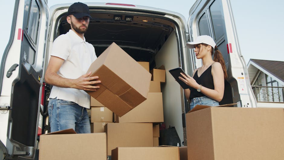 Two men are engaged in a home relocation process on a paved driveway, unloading a cardboard box with a 'Fragile' warning label from the back of a white delivery van. One man, dressed in a cap and dark jacket, is lifting the box onto a small, hand-pushed trolley, while the other, wearing glasses and a puffer jacket, assists by steadying the box. Inside the open van, there are several other cardboard boxes of various sizes, some sealed with packing tape, stacked on the vehicle's floor, indicating organised packing and furniture transport for a house move. The scene takes place outdoors near a low brick wall and a gated entrance, with natural light providing clear visibility, reflecting typical conditions during a same-day removal service. This image illustrates the logistical process of loading household items, involving careful packing materials and equipment like the trolley, as part of professional removals offered by Man and Van Crayford, focusing on efficient and secure moving and packing for house relocations.