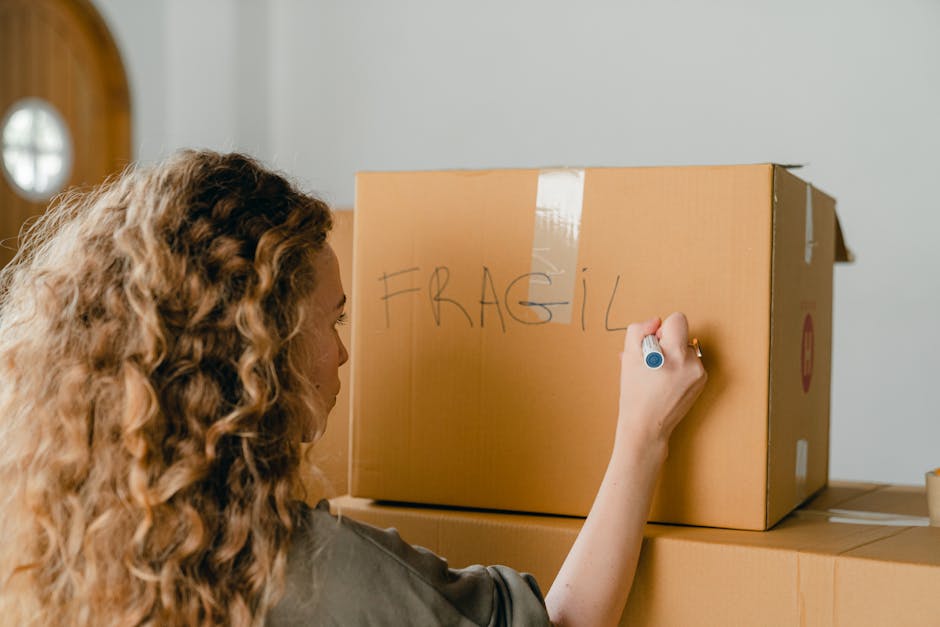 A woman with curly, light brown hair writing the word 'FRAGIL' in capital letters on a large cardboard moving box with a blue marker. The box is taped closed with packing tape and placed on top of other boxes inside a home, near a doorway with a circular window. The scene captures part of the interior environment with neutral-colored walls, reflecting a home relocation or packing process. The woman’s hand and the box are central, indicating careful handling of fragile items during a house move, with visible cardboard packaging materials and the boxed items ready for transport. This image relates to house removals and furniture transport services provided by Man and Van Crayford, focusing on packing and moving logistics as part of the home relocation process.