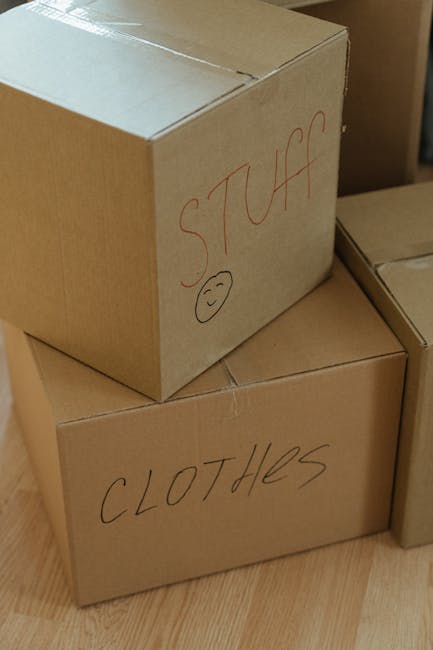 Close-up of three cardboard moving boxes stacked near a wooden floor, with the top box labeled 'STUFF' in red marker and a small smiley face drawn below. The middle box is labeled 'CLOTHES' in black marker. The boxes are sealed with packing tape and appear to be prepared for a home relocation, possibly inside a house or near an entryway. The background includes parts of other boxes and the edge of a doorframe, indicating an interior setting where packing and furniture transport preparations are underway, as seen in professional removals services like those offered by Man and Van Crayford.